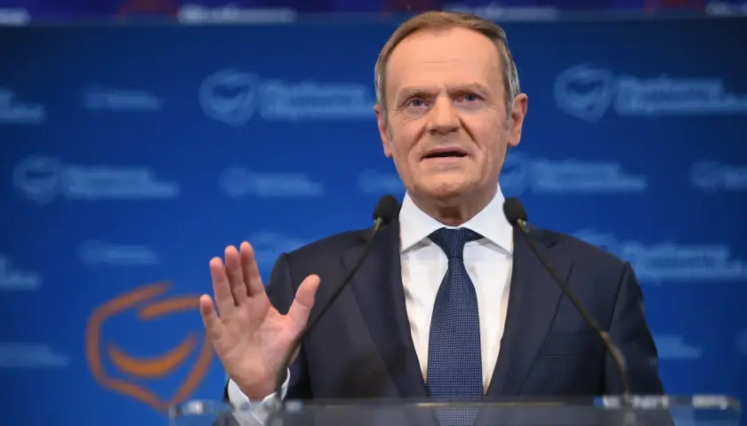 Donald Tusk w garniturze i krawacie przemawia przy mównicy z mikrofonami.
