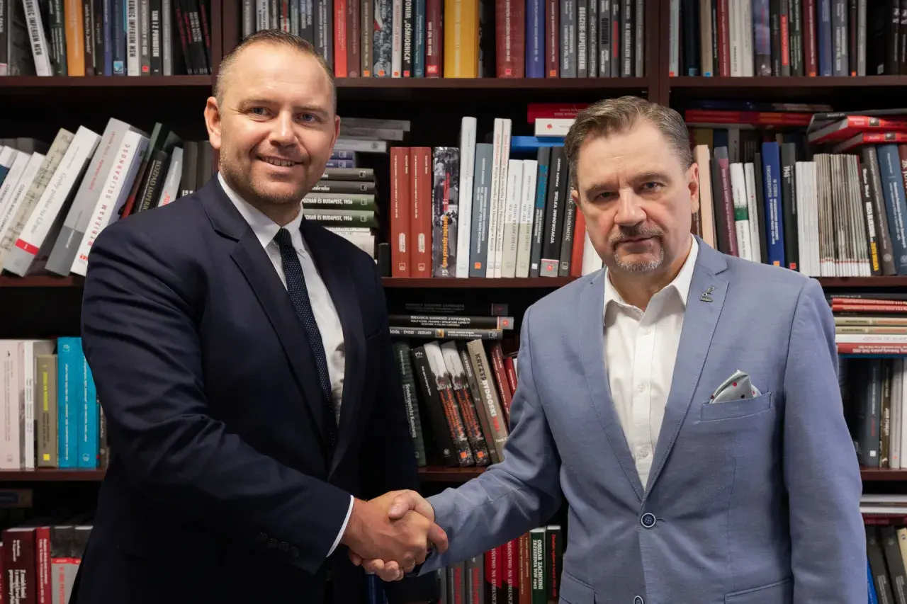 Dwóch mężczyzn w garniturach ściska sobie dłonie na tle regałów z książkami. Czy wybór Karola Nawrockiego na kandydata był konsultowany z Andrzejem Dudą?