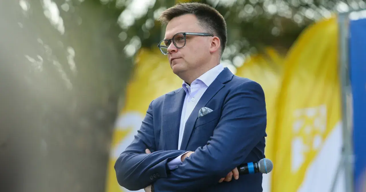 Szymon Hołownia w garniturze i okularach, z mikrofonem w ręku, na tle żółtych flag.