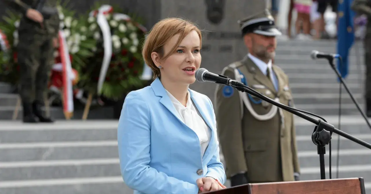 Anna Krupka w błękitnej marynarce przemawia przy mikrofonie. W tle żołnierze i wieńce. Ile milionów złotych oszczędności wykazała w oświadczeniu?