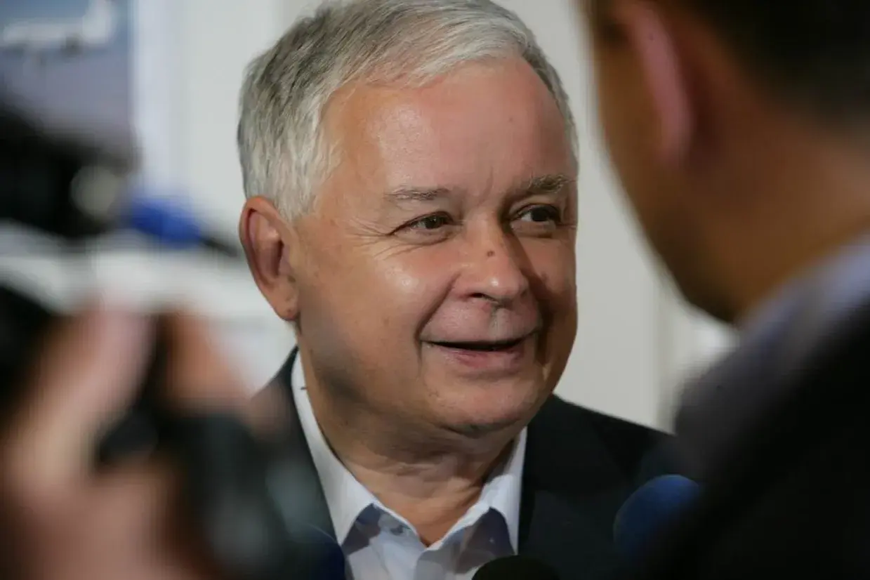 Lech Kaczyński podczas wywiadu, w tle mikrofony i niewyraźne postacie.