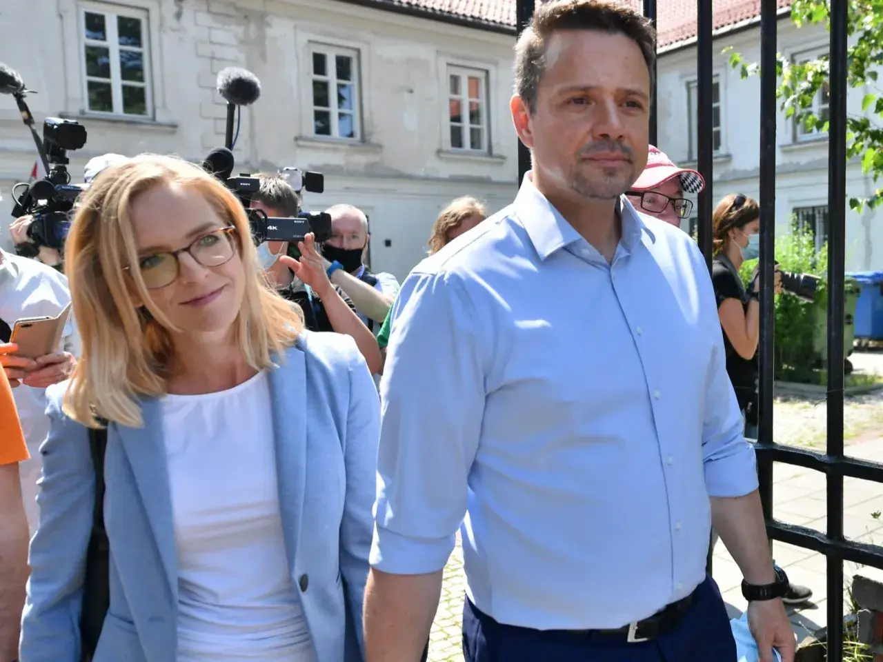 Małgorzata Trzaskowska w jasnej marynarce i Rafał Trzaskowski w błękitnej koszuli otoczeni przez dziennikarzy.
