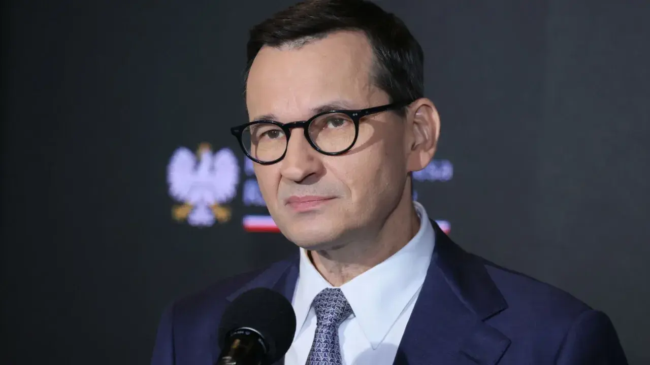 Mateusz Morawiecki w okularach i garniturze przed mikrofonem, na tle godła Polski.