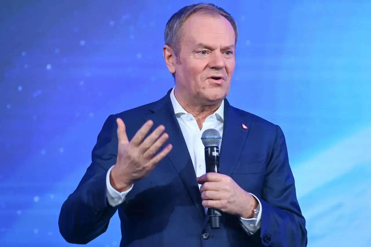 Donald Tusk w garniturze, przemawiający do mikrofonu na tle niebieskiego ekranu.