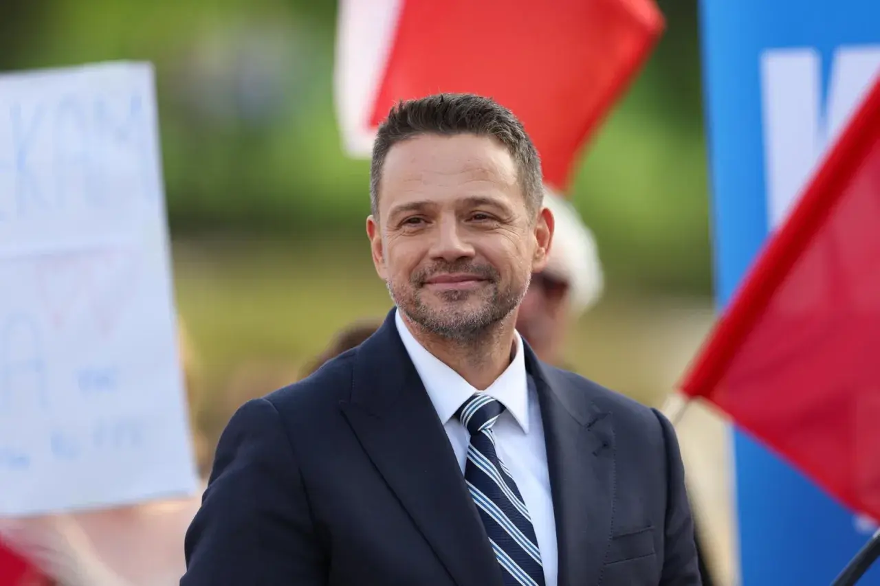 Rafał Trzaskowski w garniturze i krawacie, uśmiecha się na tle flag.