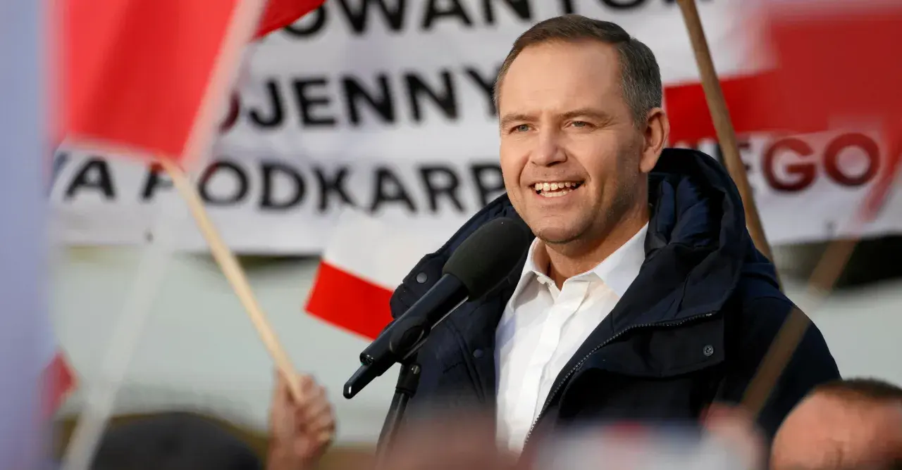 Mężczyzna w białej koszuli i granatowej kurtce przemawia do mikrofonu na tle flag.