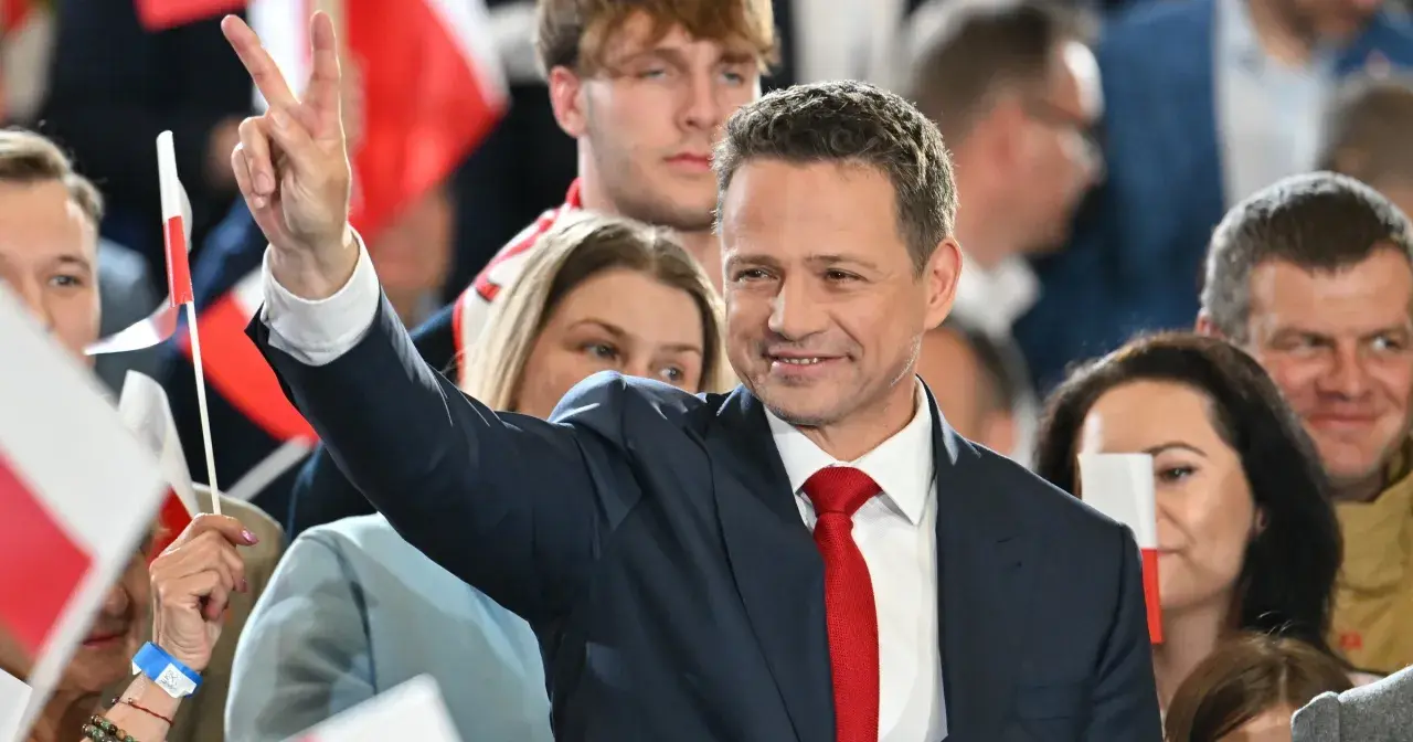 Rafał Trzaskowski w garniturze i czerwonym krawacie, pozdrawia tłum, unosząc rękę ze znakiem zwycięstwa.