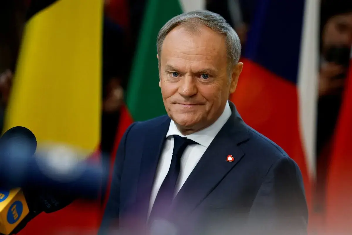 Donald Tusk, w garniturze i krawacie, z przypinką w kształcie serca, stoi na tle flag. Jego rola w Unii Europejskiej jest kluczowa.