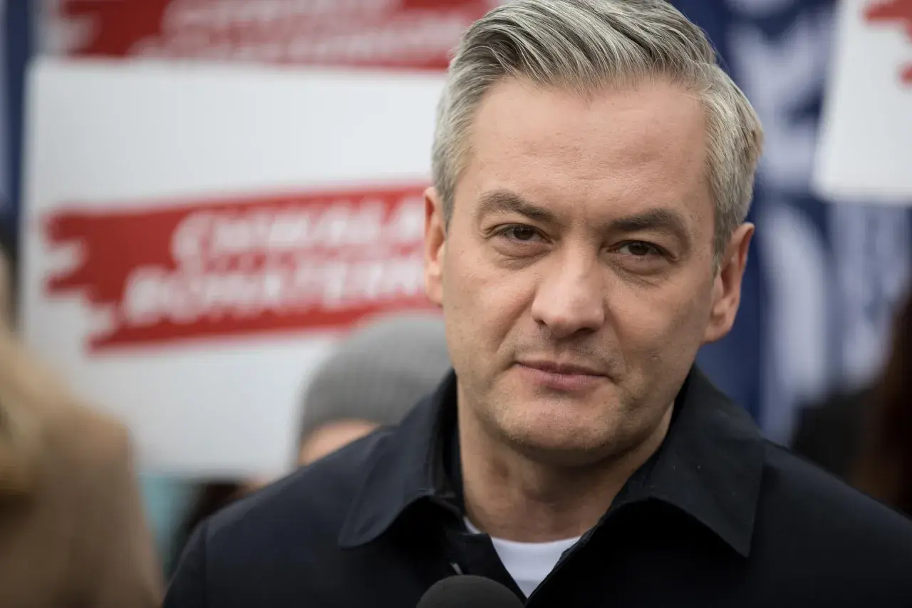 Robert Biedroń, polityk, w czarnej kurtce, z siwymi włosami. W tle czerwone napisy.