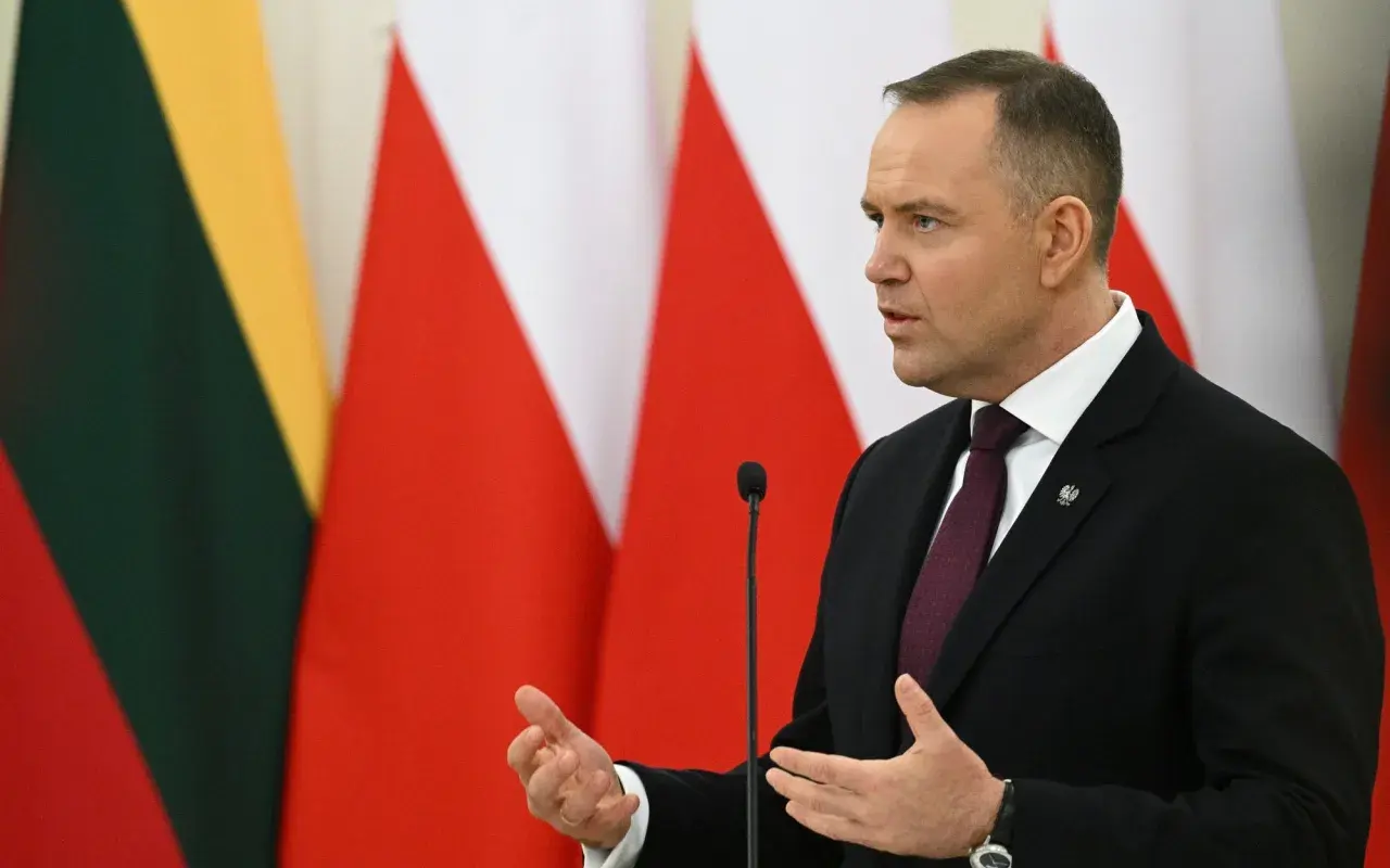 Karol Nawrocki, w garniturze, przemawia na tle flag. Jego gestykulacja i pewność siebie mogą być powodem, dlaczego Karol Nawrocki stał się nowym liderem sondaży zaufania na prawicy.