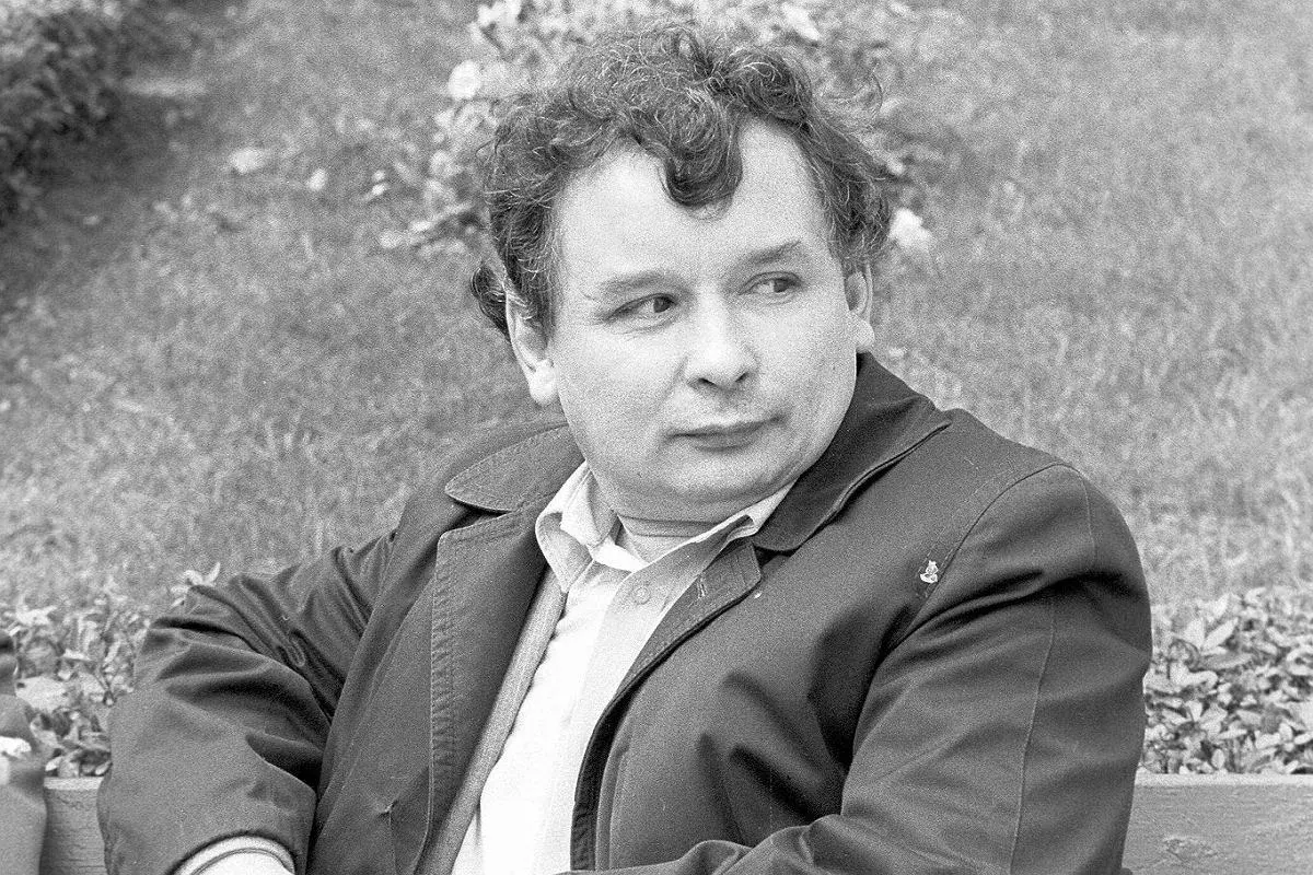 Jarosław Kaczyński w młodości, siedzący na ławce, w jasnej koszuli i ciemnej kurtce.
