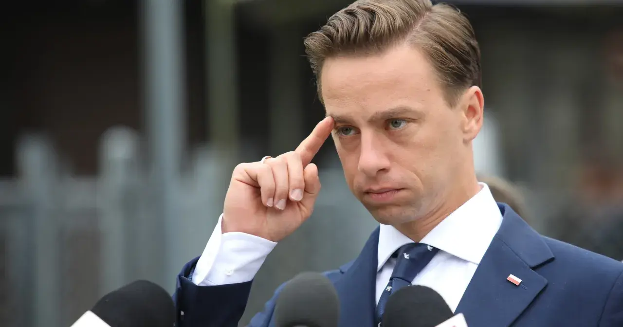 Krzysztof Bosak w garniturze, z palcem przy skroni, przed mikrofonami.