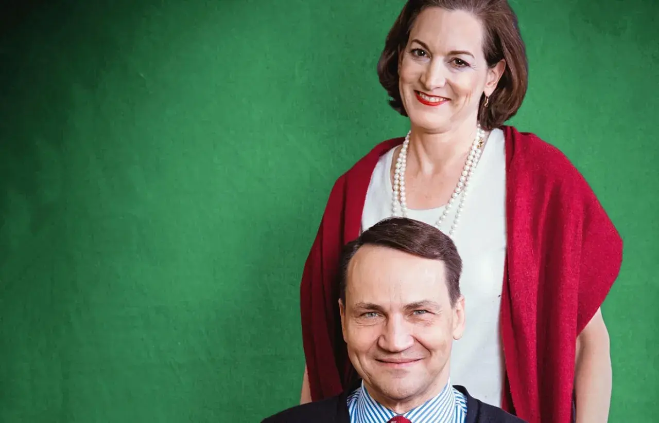 Uśmiechnięta kobieta w perłach i czerwonym szalu stoi za mężczyzną w pasiastym krawacie.