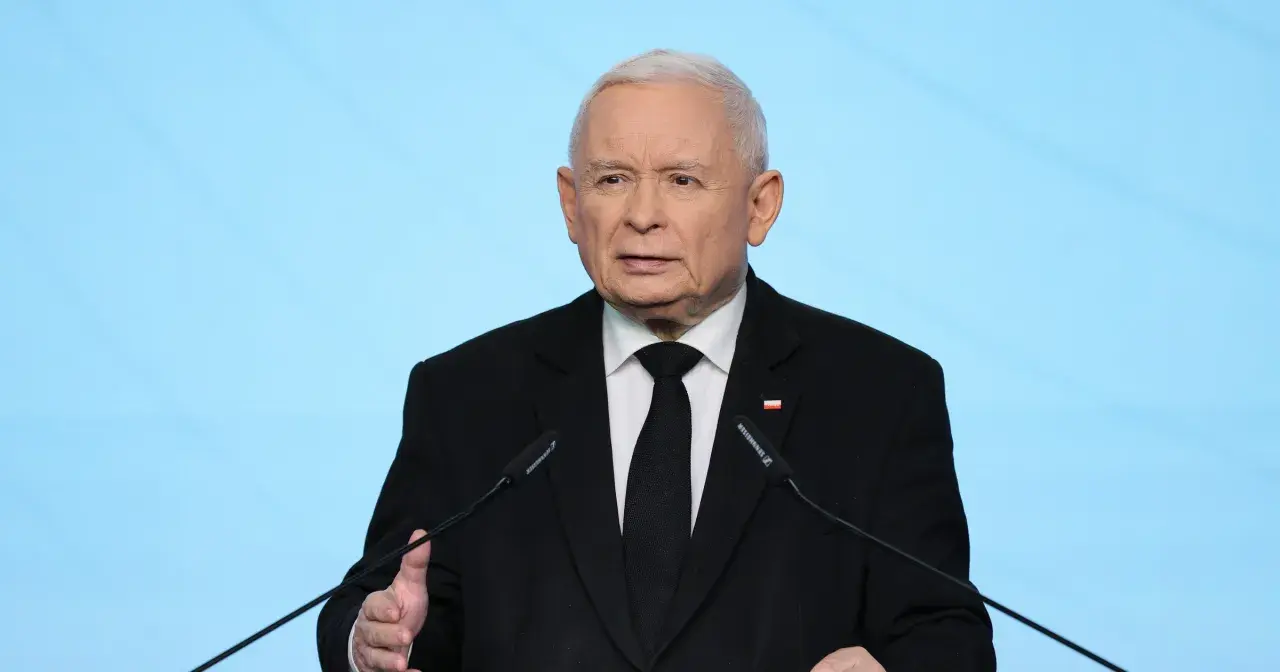 Jarosław Kaczyński w czarnym garniturze przemawia przy mikrofonie na tle niebieskiego tła.
