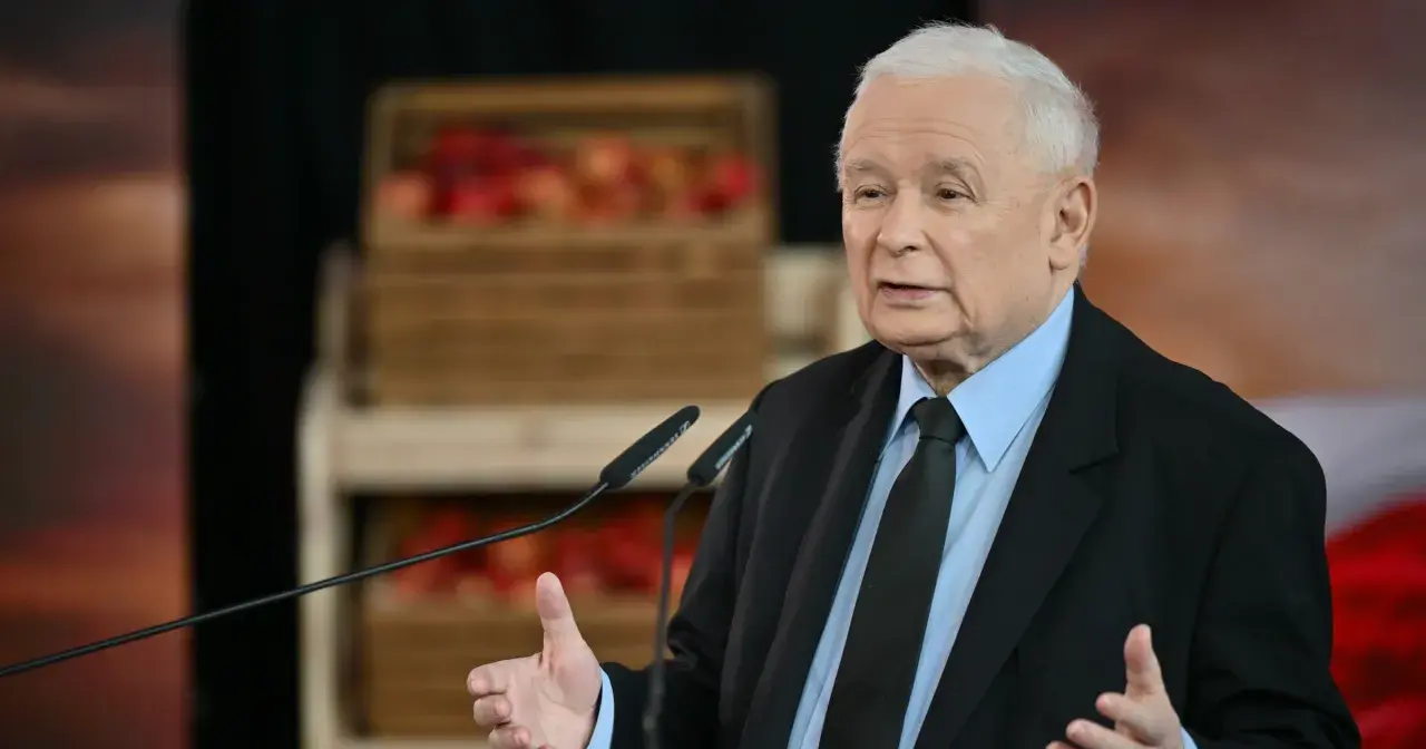 Jarosław Kaczyński w garniturze przemawia przy mikrofonach, w tle skrzynki z jabłkami.