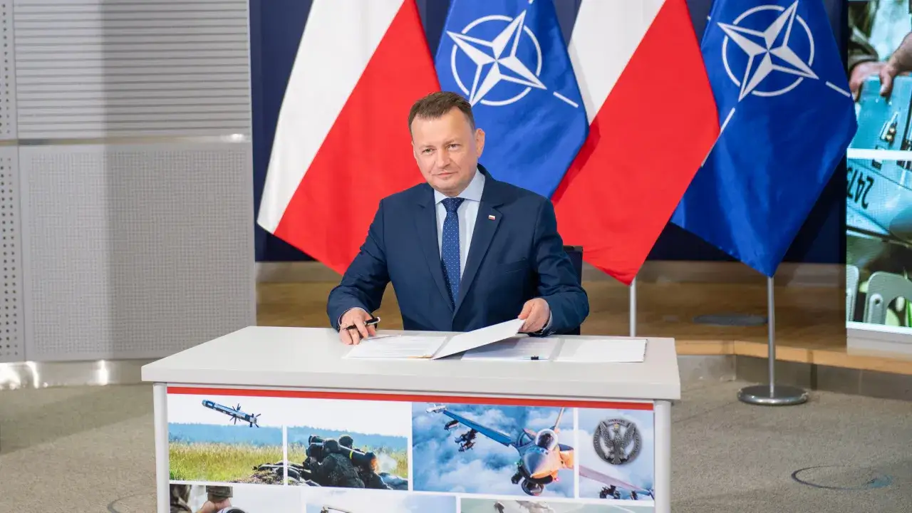 Minister podpisuje dokumenty, za nim flagi Polski i NATO. Na stole grafiki wojskowe, sugerujące sekrety funduszu bezpieczeństwa i obronności ukryte w ustawie o administracji.