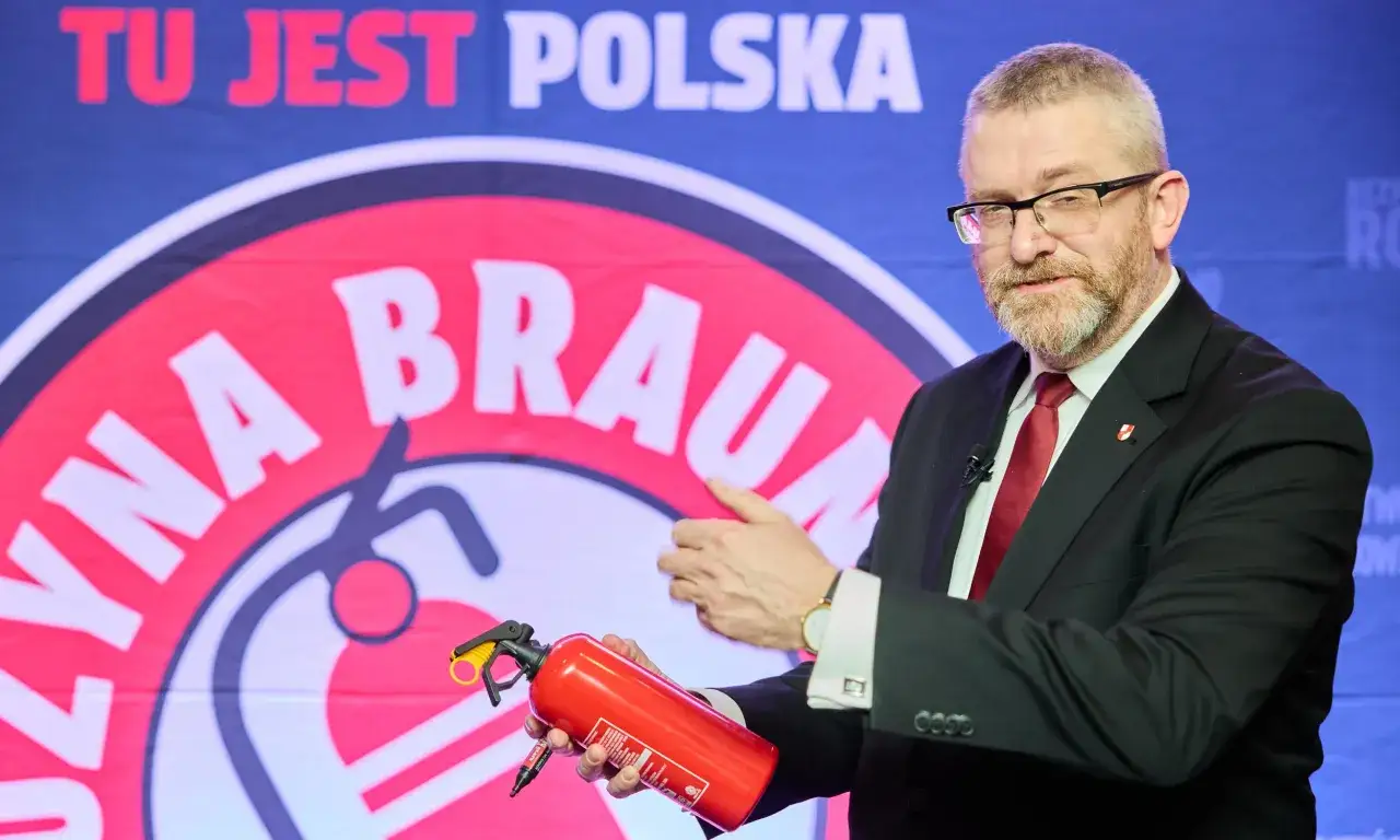 Grzegorz Braun w garniturze trzymający gaśnicę na tle banera z napisem "TU JEST POLSKA".