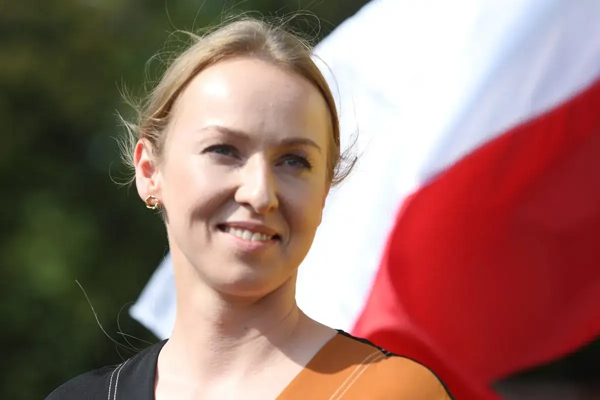 Uśmiechnięta Urszula Brzezińska Hołownia, z polską flagą w tle, symbolizuje dumę z tego, skąd pochodzi.