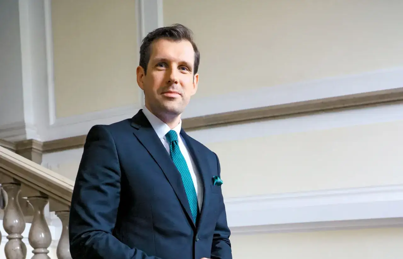 Tobiasz Bocheński w garniturze, z zielonym krawatem i poszetką, stoi na tle eleganckich schodów. To może być odpowiedź na pytanie, dlaczego Tobiasz Bocheński jest promowany na nową twarz polskiej prawicy.