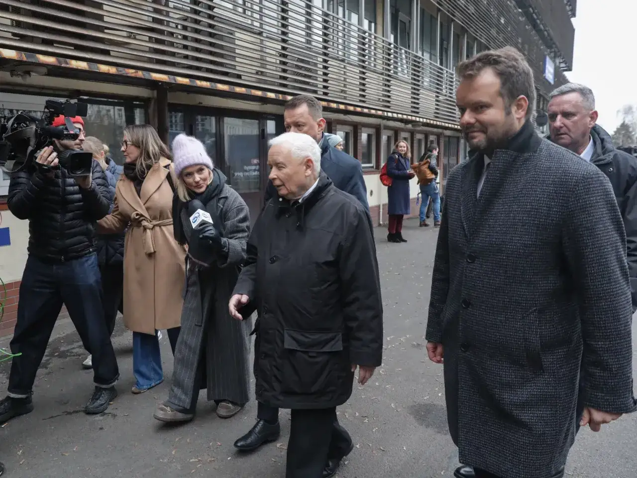 Jarosław Kaczyński w otoczeniu współpracowników, w tym kobiety z mikrofonem TVN, idzie z innymi politykami.