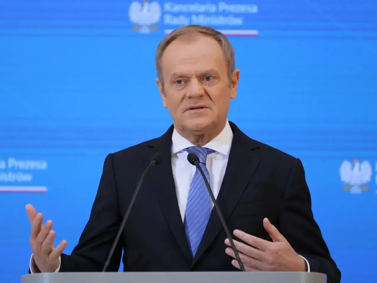 Czy Donald Tusk osobiście interweniował w sprawie blokady drogi Jasło-Krosno w lutym? Polityk przemawia na tle niebieskiej ściany z godłem Polski.
