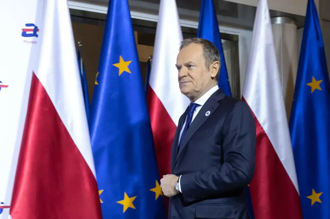 Donald Tusk, w garniturze, stoi przed flagami Polski i Unii Europejskiej. Jego obecność podkreśla, jaką funkcję pełni Donald Tusk w Unii Europejskiej.