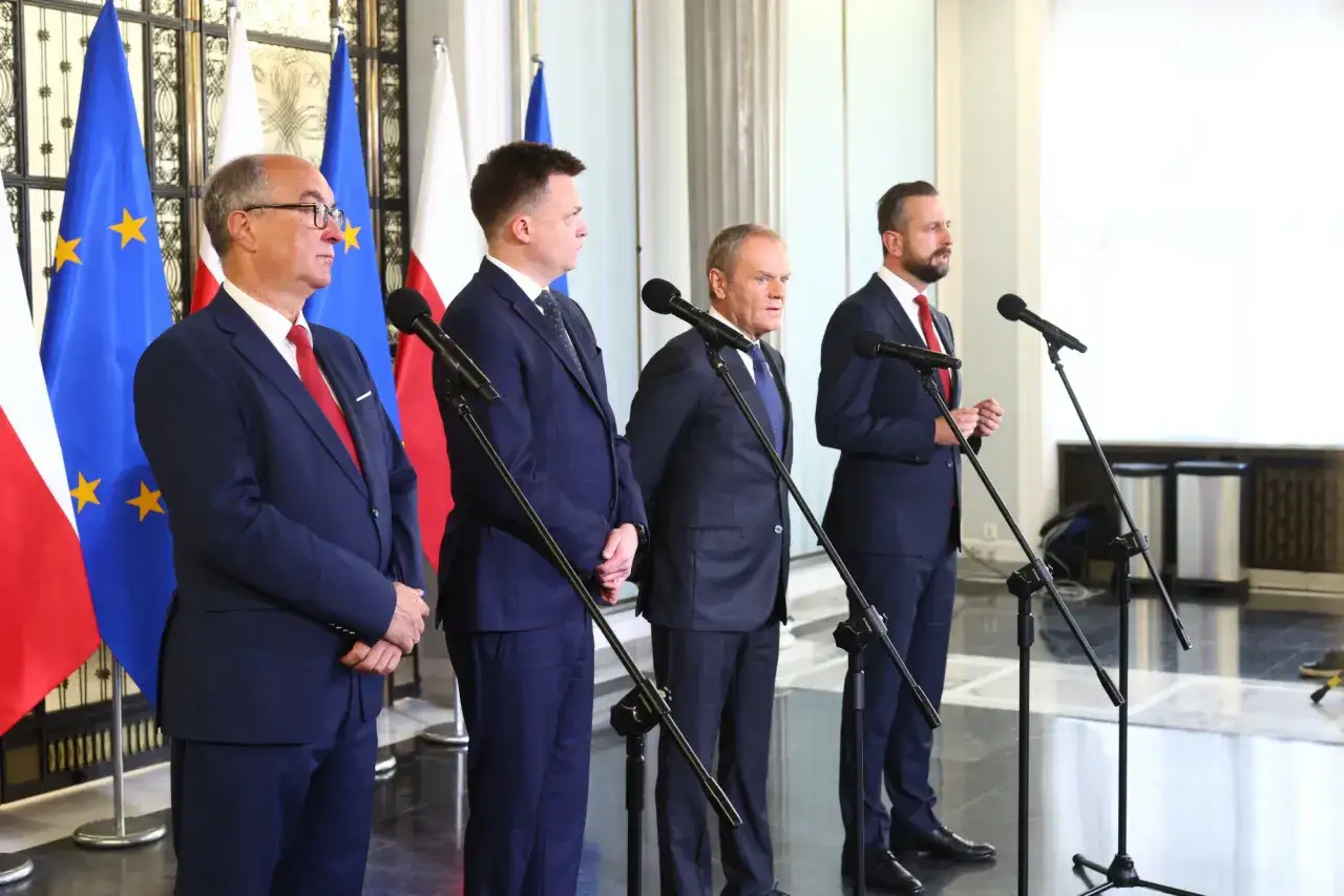 Politycy w garniturach stoją przed flagami. Dyskusja o tym, dlaczego kandydatura Szymona Hołowni na prezydenta budzi sprzeciw wewnątrz koalicji rządzącej.