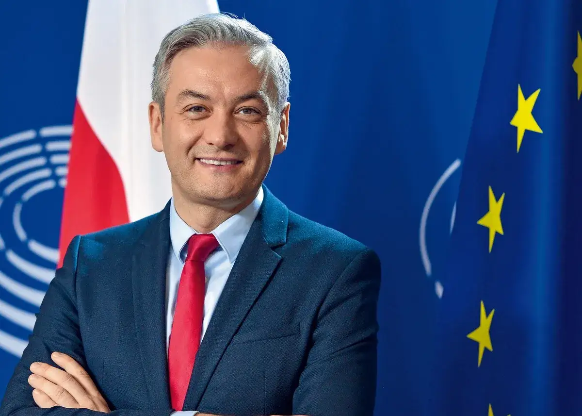 Robert Biedroń, polityk, uśmiecha się na tle flagi Polski i UE. Ile lat ma Robert Biedroń?