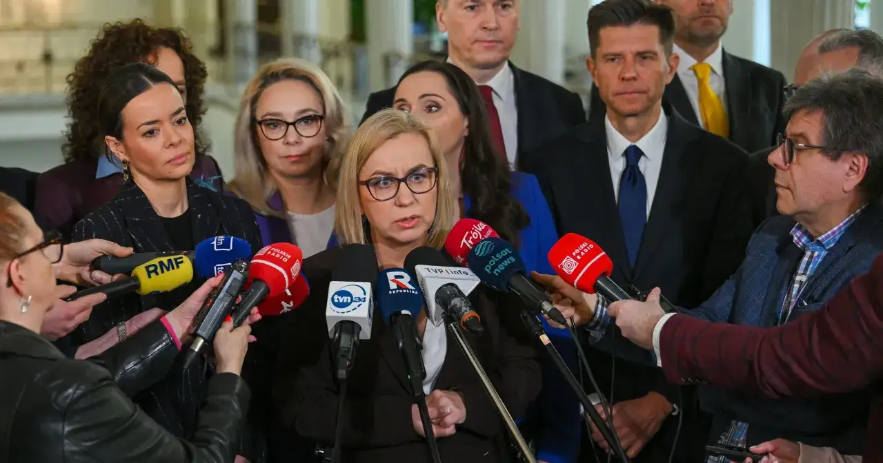 Kto stoi za nowym programem partii? Kobieta w okularach, otoczona przez dziennikarzy z mikrofonami, udziela wywiadu.