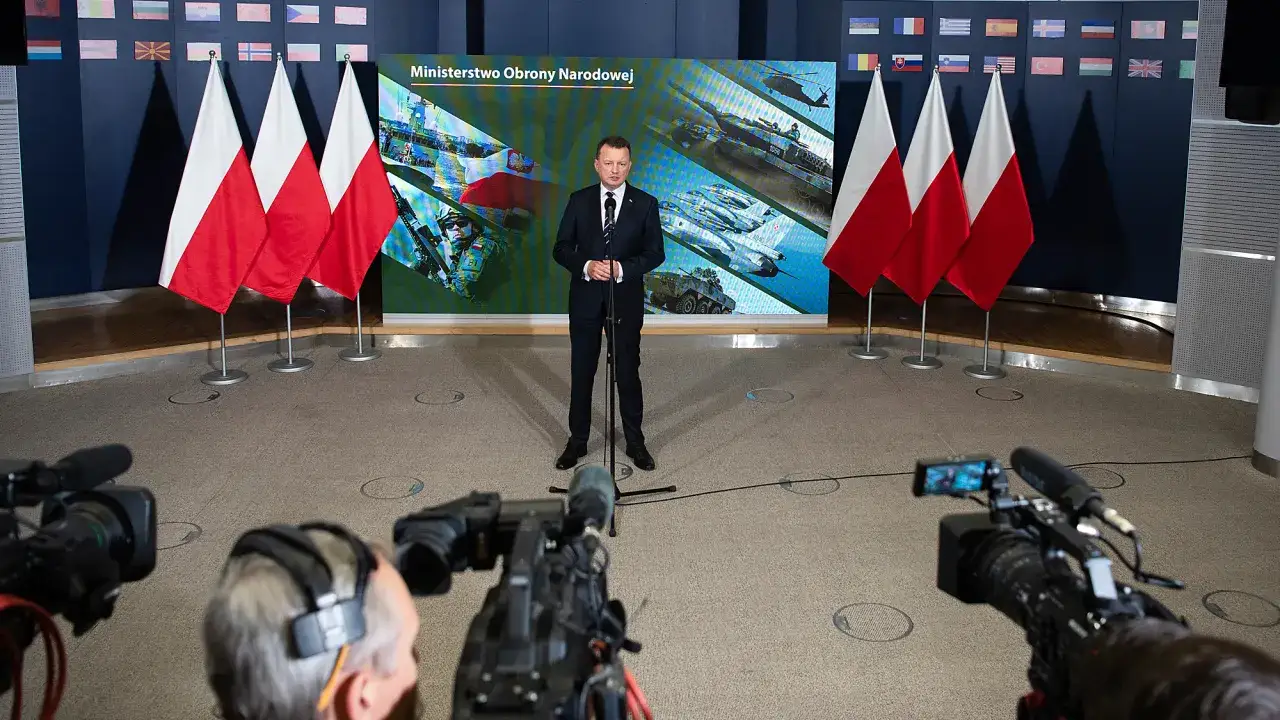 Minister przemawia, ujawniając sekrety funduszu bezpieczeństwa i obronności ukryte w ustawie o administracji. W tle flagi i ekrany z grafikami wojskowymi.