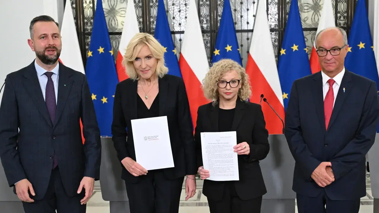Cztery osoby stoją przed flagami Polski i UE. Dwie kobiety trzymają dokumenty, co może być związane z tym, dlaczego koalicja rządząca spiera się o ustawę dotyczącą związków partnerskich.